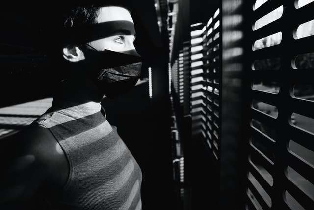 woman in a dark room with a dark mask looking through blinds at the light outdoors